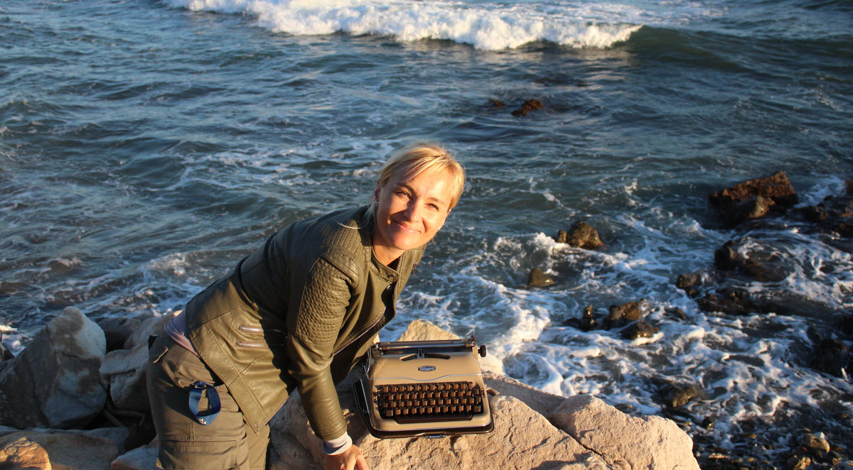 A woman with blonde hair sits on a rock by the ocean, smiling at the camera. A vintage-style typewriter rests on the rock in front of her.