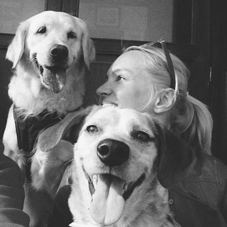 A black and white photograph showing a woman smiling while hugging two dogs, one golden retriever and one mixed breed, with both dogs panting happily.