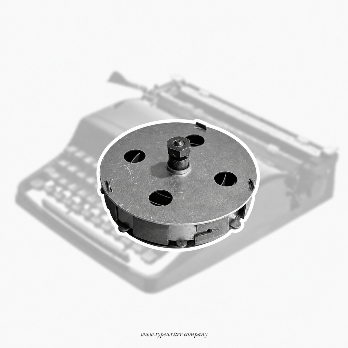 A circular metal typewriter drum or roller component shown in front of a faded, grayscale background of a vintage typewriter. The product is isolated with a white outline on a light gray background.
