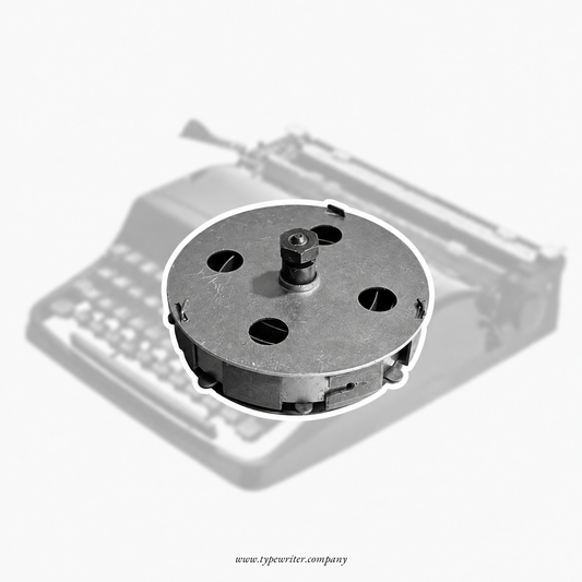 A circular metal typewriter drum or roller component shown in front of a faded, grayscale background of a vintage typewriter. The product is isolated with a white outline on a light gray background.