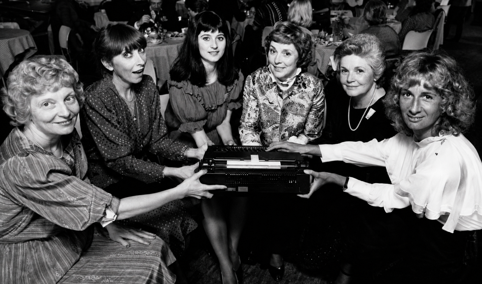Black and white photograph of six women gathered together in a social setting, holding a rectangular object in the center.
